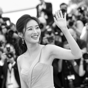 Nam Gyu-Ri, Asian actress, waving and smiling at the Cannes Film Festival. Black and white halter neck dress. Background is a sea of flashing cameras and enthusiastic fans.