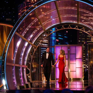 John C. McGinley and Nichole Kessler at a glamorous awards show. Both dressed in designer attire. John in a sharp black tuxedo. Nichole in a form-fitting red dress. The backdrop is a sparkling city skyline.