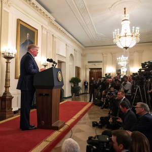 Donald Trump at a press conference, standing behind a podium in the East Room of the White House.