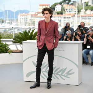 Timothée Chalamet, standing in a green room at the Cannes Film Festival. Wearing a maroon blazer and black pants. Surrounded by photographers and festival attendees