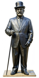 A statue of Hercule Poirot as played by David Suchet, a bowler hat on his head, fine mustache, hands in gloves, with a walking cane