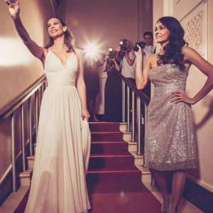 Sinta Weisz and Shermine Sharivar at a glamorous awards ceremony. Sinta in a white gown with a deep V-neckline. Shermine in a sparkling silver dress. The backdrop is a sea of flashing cameras and excited fans.