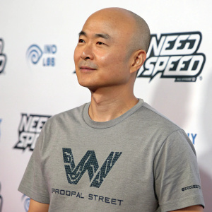 C.S. Lee, an Asian man with a bald head, is photographed against a white backdrop at a gaming convention. He wears a casual gray t-shirt with a W-shaped logo from Need for Speed ProStreet.