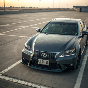 A sleek, dark grey Lexus GS 350 parked in an empty lot. The car's prominent logo and mesh-like grille pattern are clearly visible. Contemporary design with a license plate showing the model code. Lighting highlights the car's refined lines and shape.