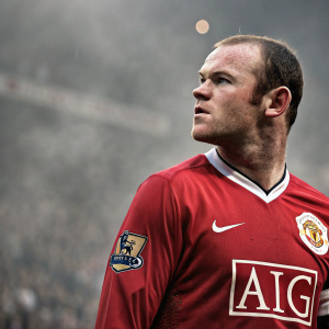 Wayne Rooney, intense gaze, wearing Manchester United jersey with AIG logo. Faint crowd in background.
