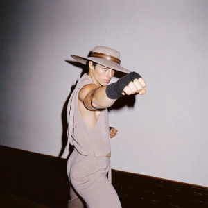 Kung Lao from Mortal Kombat, striking a fighting pose. Wearing a snug beige outfit and a wide-brimmed hat. Dark, shadowy backdrop