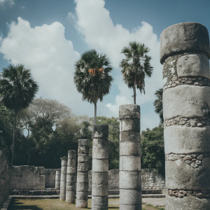 Ancient stone columns at Chichen Itza, a UNESCO World Heritage Site in Mexico. Weathered stone, rows of columns, lush greenery, tall palm trees. Vibrant blue sky with scattered clouds. Pillars are the main focus, surrounded by rich textures and natural elements.