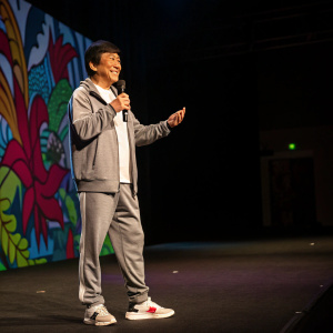 Jackie Chan, in a casual sporty outfit, stands on a dimly lit stage. He poses dynamically with a confident smile. A colorful backdrop is behind him.