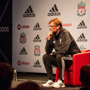 Jürgen Klopp, seated on stage, during a Liverpool FC press conference. Backdrop features logos of Adidas and EA Sports. Candid moment with a thoughtful expression.