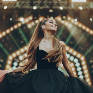 Ariana Grande, with long flowing hair, elegant black dress, standing on vibrant stage backdrop.