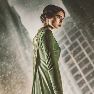 Margaret Qualley, intense expression, dark brown hair styled elegantly. Wearing a green dress with gold detailing. The background is a blurred cityscape.