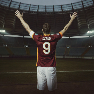 Edin Dzeko, soccer star, mid-goal celebration. Arms raised high in the air. His maroon and gold jersey with orange accents clearly visible. The name 'DZECO' and number 9 on the back. A vast stadium with sparse audience and a pristine grass field in the background