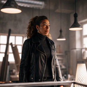 Tawny Cypress as Nez Rowan in 'The Blacklist: Redemption'. Wearing black leather jacket and dark turtleneck. Stands in a brightly lit studio with modern art installations in the background.