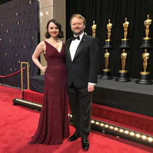 Karina Longworth, dressed in an elegant maroon gown, and Rian Johnson, in a classic black tuxedo, at the Critics' Choice Awards. The stage is set with gold trophies and red carpet.
