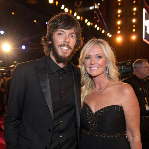 Chris Janson and Kelly Lynn, dressed in formal attire, at the ACM Honors gala. Bright lights and cameras in the background.
