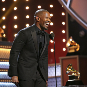 Jamie Foxx at the Grammy Awards. Bald head, black suit, bow tie, grinning widely. Stage filled with awards and bright lights.
