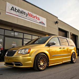 Shiny gold Chrysler Town and Country minivan parked outside AbilityWorks building. Bold company logo visible on banner.