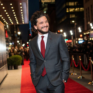 Kit Harington, wearing a stylish charcoal suit and red tie, at the grand opening of a new art gallery. Bright city lights and bustling crowd in the background.