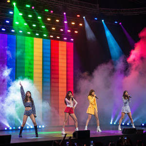 Korean girl group 4Minute performing under vibrant LED lights and dramatic stage effects. Multi-colored backdrop, members in various outfits and poses, showcasing their stage presence.