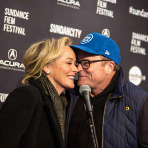 Sharon Stone and Tom Arnold sharing a heartfelt moment at the Sundance Film Festival. Stone has blonde hair, while Arnold sports a blue cap and glasses. They are standing close together with a microphone in front of them, against a dark backdrop with festival logos.
