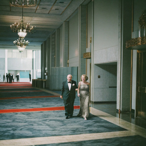 Ed Asner and Liza Asner attending a gala event. They both look elegant in formal attire. The venue is adorned with chandeliers and red carpets.