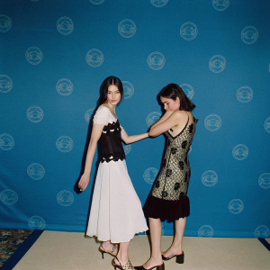 Margaret Qualley and Rainey Qualley attending a sustainable fashion event. Margaret in a black and white ensemble with a white skirt, Rainey in a black and gold lace dress. The backdrop features eco-friendly logos.