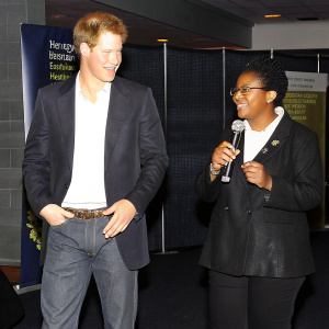 Prince Harry and Yvette Twagiramariya at a health fair. Standing in front of banners promoting HIV awareness and testing initiatives. Indoor setting with soft lighting and modern backdrop.