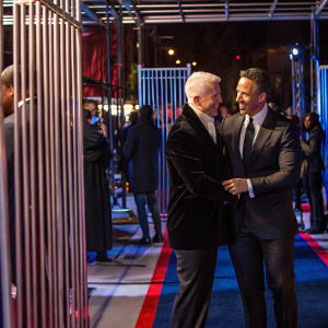 Anderson Cooper and Chris Cuomo at the annual WarnerMedia event. Both in sharp black suits, they embrace warmly. White backdrop with WarnerMedia logos. Blue carpet event setting.