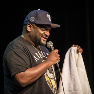 A close-up of comedian Aries Spears. He wears a Yankees cap and graphic t-shirt, holding a white towel. Onstage with microphone. Dark backdrop