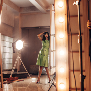 Krysten Ritter on the set of a fashion photoshoot. She's in a vibrant green dress, posing confidently. Studio lights, sleek black background