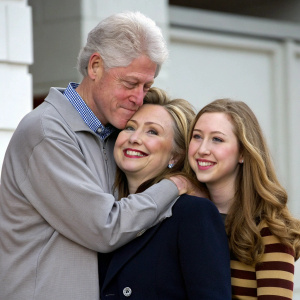 A family photo of Bill Clinton, Hillary Clinton, and Chelsea Clinton. Bill embraces Hillary while Chelsea stands nearby.