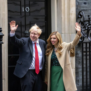Boris Johnson and Carrie Symonds waving from 10 Downing Street. Boris in dark suit with red tie, Carrie in beige coat and green skirt. Behind them, black ironwork and the iconic black door