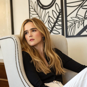 Close-up shot of Zoey Deutch. She has long blonde hair and minimal makeup. Sitting in a cozy armchair with modern black and white wall art behind her.