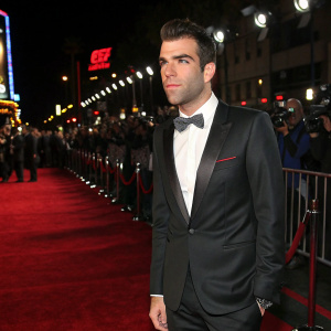 Zachary Quinto on a glamorous Hollywood premiere. Wearing a sleek black suit, white shirt, and bow tie. Glowing red carpet, flashing cameras.