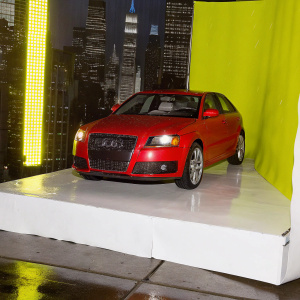 A shiny red Audi RS3 parked in front of a neon-lit cityscape at night. The car's athletic design and LED headlights stand out against the vibrant urban backdrop.