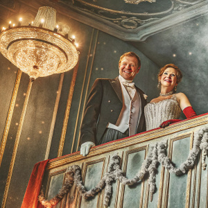 Carl Eduard Graf von Bismarck and Countess Nathalie von Bismarck, grinning at a lavish ballroom event. Grand chandelier hanging above, intricate decor