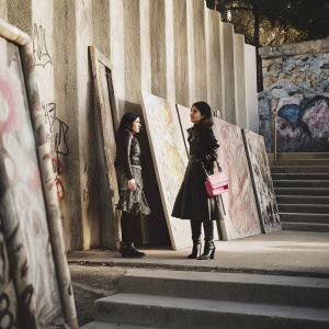 Isabelle Fuhrman and Elina Fuhrman at an outdoor art gallery. The backdrop features vibrant graffiti and street art. Isabelle in a chic leather dress with a pink handbag. Elina in a stylish black layered outfit.