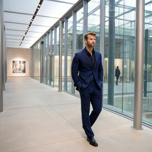 Glen Powell at a sleek, modern gallery opening. Wearing a dark blue suit. Indoor setting with glass walls and steel columns.