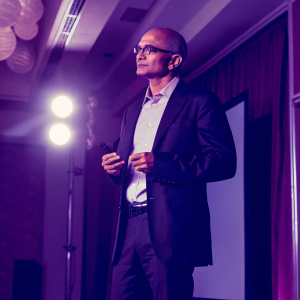 Satya Nadella, Microsoft CEO, standing on a stage during a tech conference. Bright lights highlight him. He wears a dark suit, white shirt, and glasses. Serious expression. Large screen displays keynotes behind him.