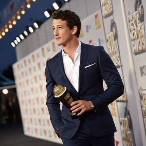 Miles Teller at the MTV Movie Awards. Dressed in a navy suit and white shirt. He is holding an award. The backdrop features intricate award details.