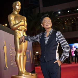 Raymond Wong Pak-Ming, an Asian film producer and actor, poses with a large golden statue at a gala event. Wearing a stylish striped shirt and dark vest.