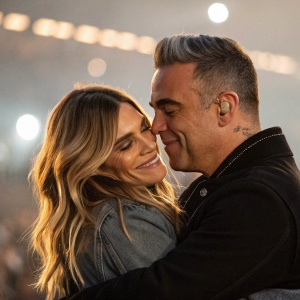 Robbie Williams and Ayda Field, a blissful couple, sharing a warm hug. Soft ambient lighting, blurred crowd in background. Candid photo capturing genuine affection.