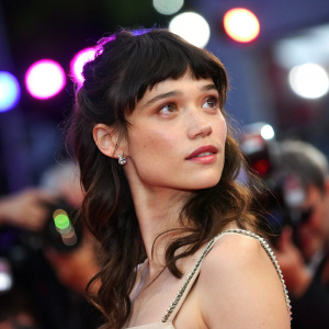 Close-up of Astrid Berges-Frisbey at Cannes Film Festival. She has a wistful look. Dark hair with bangs. Background is a mix of bright colors and lights.