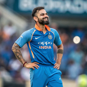 Virat Kohli, bearded, in blue cricket jersey with 'oppo India' logo. Orange accents, standing in stadium. Confident smile, tattoos visible on arms. Background blurred.