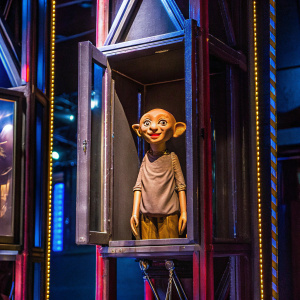 A Dobby doll from the Harry Potter series, in original packaging. White box with Harry Potter branding. Playful expression with wide eyes, dressed in earth-toned outfit. Interactive elements included. Photo style: realistic