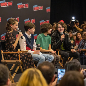 Natalia Dyer, Noah Schnapp, Finn Wolfhard, and Millie Bobby Brown seated on chairs during a Q&A session at New York Comic Con. Crowded room with fans in the background.
