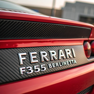 Close-up of a sleek red Ferrari F355 Berlinetta. The car's nameplate, in silver text, stands out against the textured carbon fiber material. The blurred background highlights the vibrant red paint and intricate design.