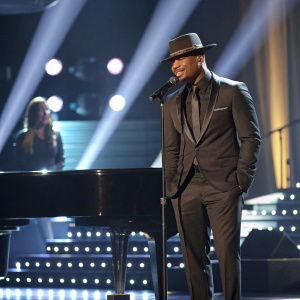 Ne-Yo, stylish in a black suit and fedora, performs at the Grammy Awards. The stage is filled with lights and a grand piano.