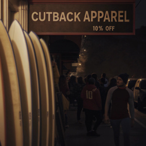 Cutback Apparel grand opening celebration on a bustling urban street. Surfboards displayed prominently, 10% off special offer. Crowded with shoppers and event-goers