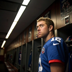 Colt McCoy, intense stare, NY Giants jersey, indoor locker room, posters and equipment visible.
